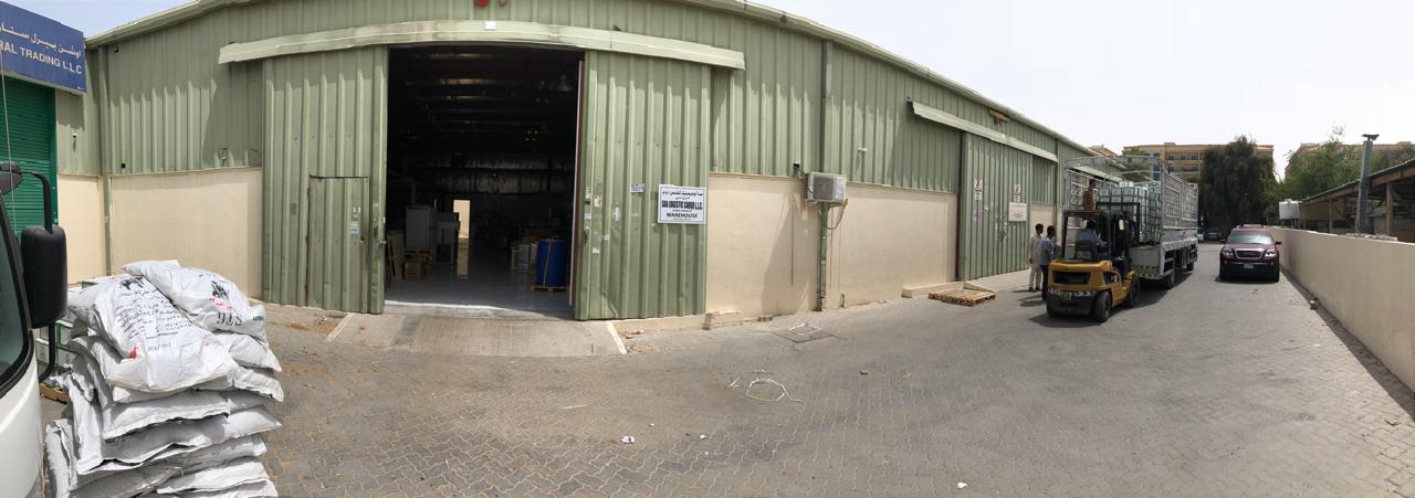 Panoramic view of SDA Logistics Services Company's warehouse exterior with an open bay door, a forklift in motion, and various goods awaiting shipment.