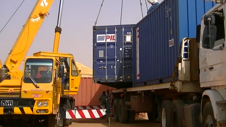 A yellow crane lifting a blue shipping container onto a truck at a logistics hub.